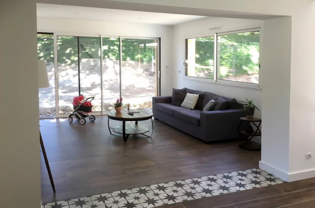 Un salon lumineux et spacieux grâce à une extension bien pensée. Les baies vitrées panoramiques apportent une vue imprenable sur l'extérieur tout en baignant la pièce de lumière naturelle. Le parquet effet bois donne une ambiance chaleureuse, contrastée par une touche d'originalité avec le carrelage graphique. Un aménagement moderne et accueillant, parfait pour les moments de détente en famille.
