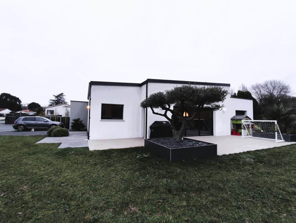 Maison de style contemporain avec une façade blanche, une terrasse spacieuse ornée d'un olivier dans un bac moderne, et un aménagement extérieur comprenant un espace de jeux pour enfants et une pelouse bien entretenue.
