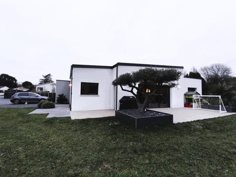 Maison de style contemporain avec une façade blanche, une terrasse spacieuse ornée d'un olivier dans un bac moderne, et un aménagement extérieur comprenant un espace de jeux pour enfants et une pelouse bien entretenue.