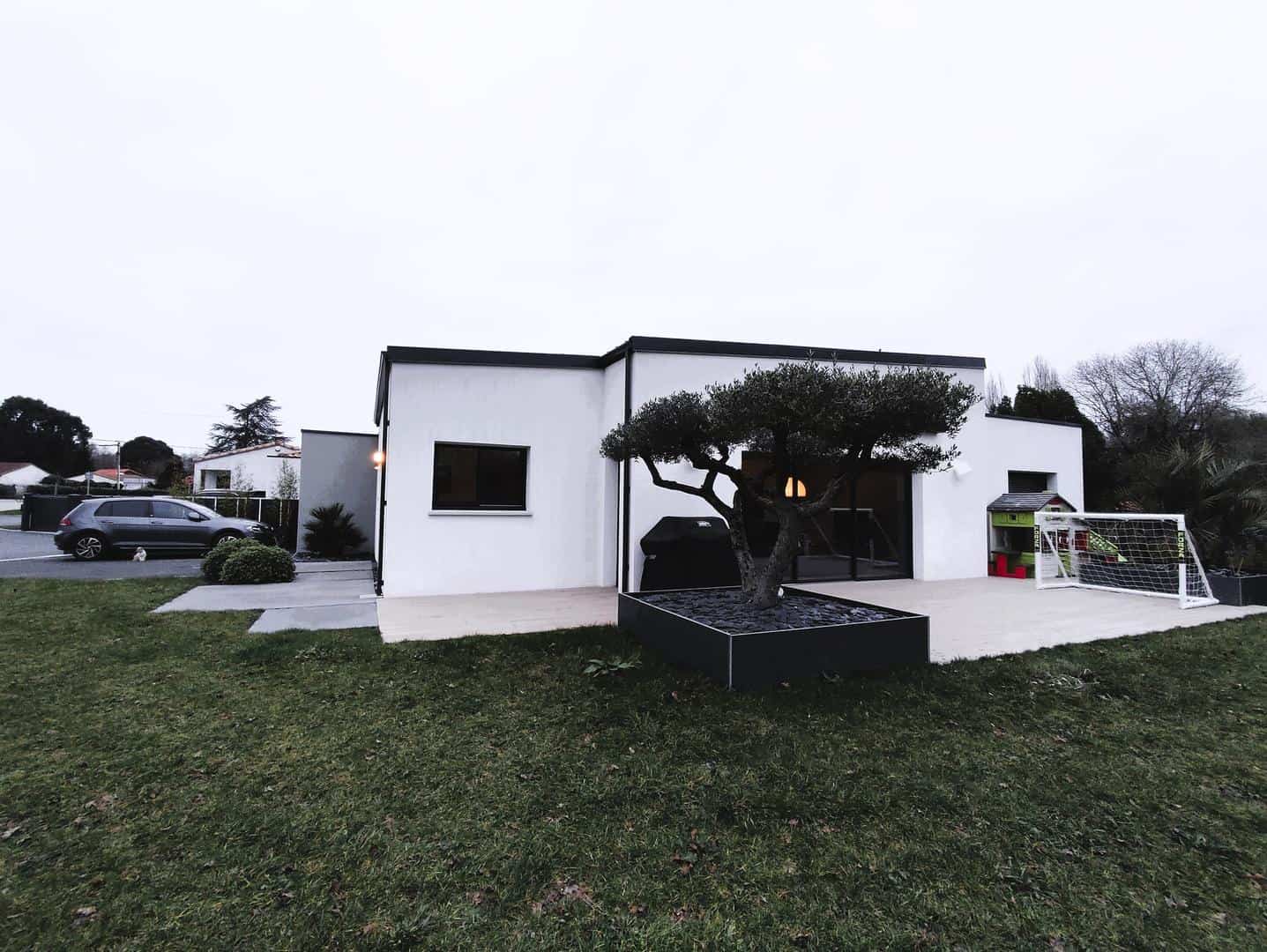 Maison de style contemporain avec une façade blanche, une terrasse spacieuse ornée d'un olivier dans un bac moderne, et un aménagement extérieur comprenant un espace de jeux pour enfants et une pelouse bien entretenue.