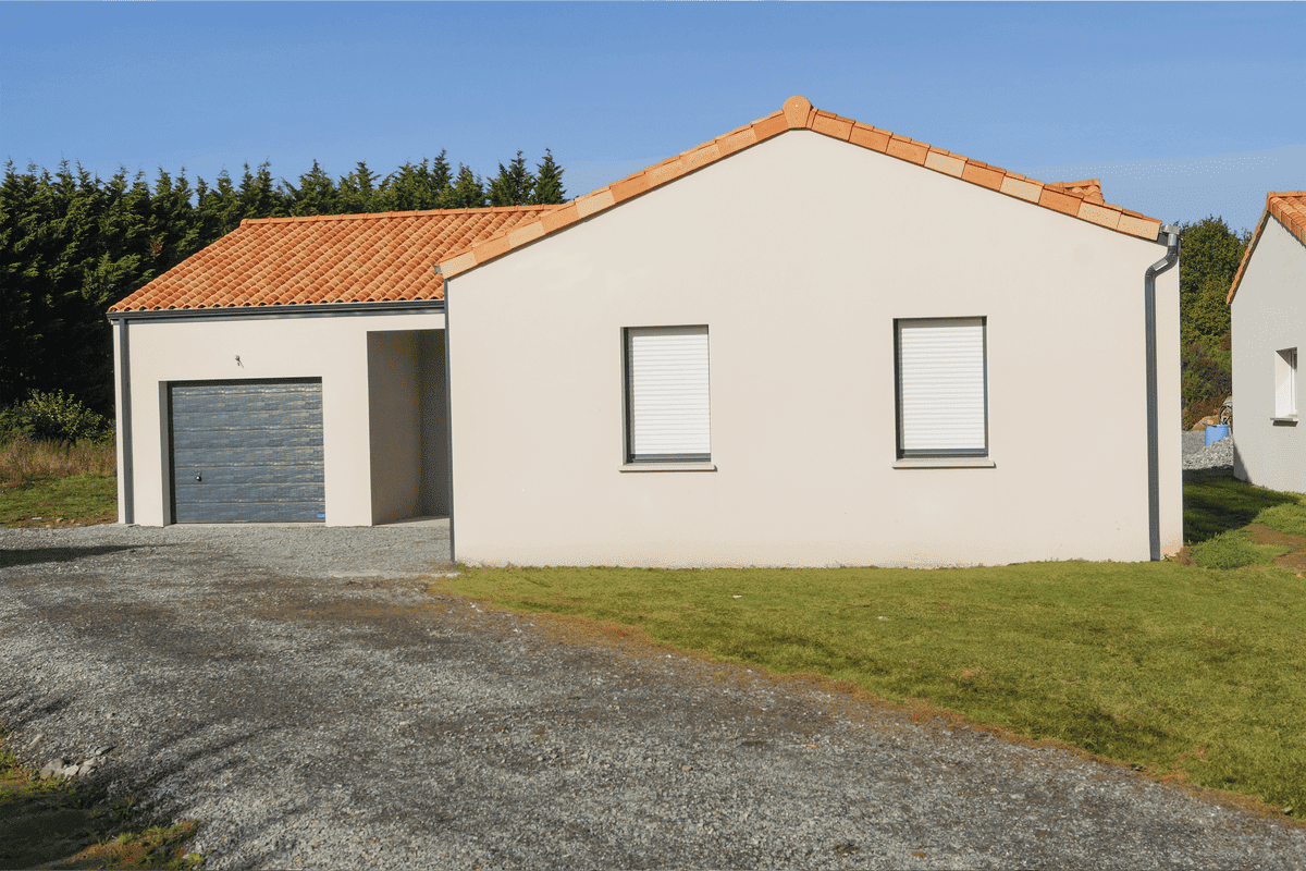 Maison de plain-pied en crépi beige avec toit en tuiles rouges, garage intégré et allée en gravier sur terrain arboré.