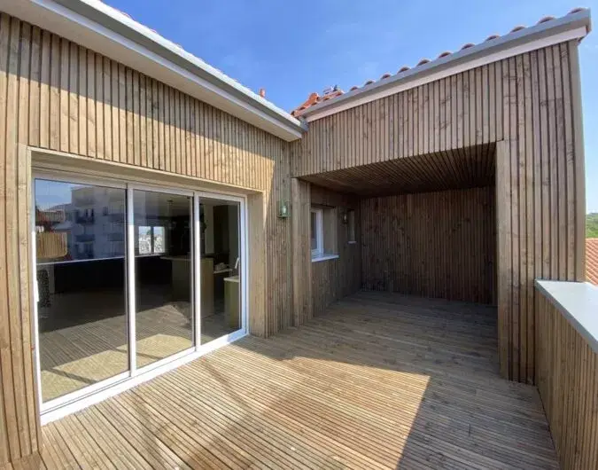 Une terrasse en bois élégante et contemporaine, idéale pour profiter des journées ensoleillées. Le revêtement en bois naturel offre une atmosphère chaleureuse et harmonieuse, parfaitement intégrée à l'architecture de la maison. L'espace semi-couvert ajoute une touche de praticité, permettant une utilisation en toute saison. Une extension extérieure qui allie confort et esthétisme.