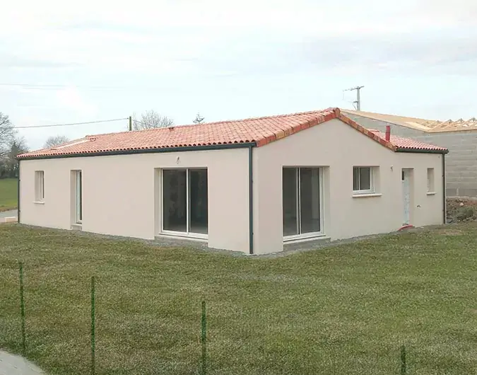 Maison de plain-pied avec toit en tuiles rouges, façade beige et grandes baies vitrées, située sur un terrain herbeux, sous un ciel nuageux.