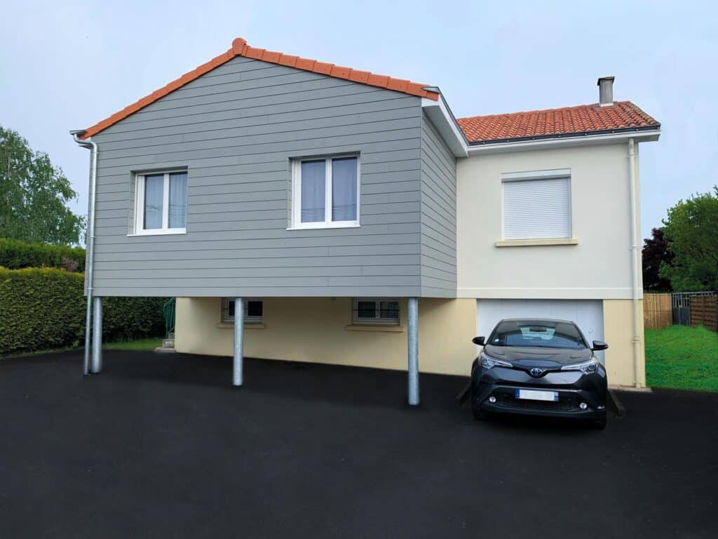 Maison avec une extension en bardage gris, voiture stationnée devant, et sous-sol sur pilotis.