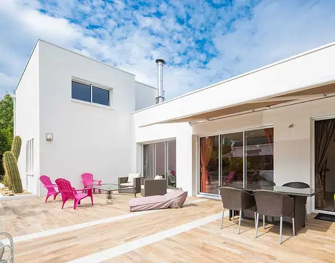 Maison contemporaine avec grande terrasse en bois, mobilier de jardin coloré, et baies vitrées, sous un ciel partiellement nuageux.