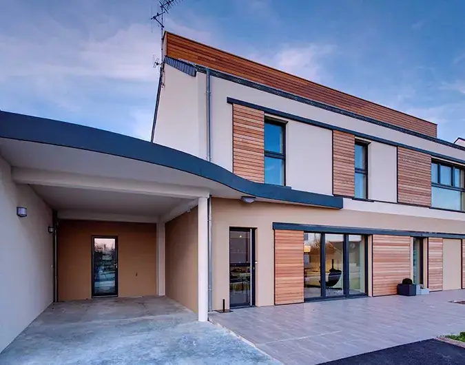 Maison passive à deux étages avec bardage en bois et grandes baies vitrées, conçue pour une haute performance énergétique, sous un ciel crépusculaire.