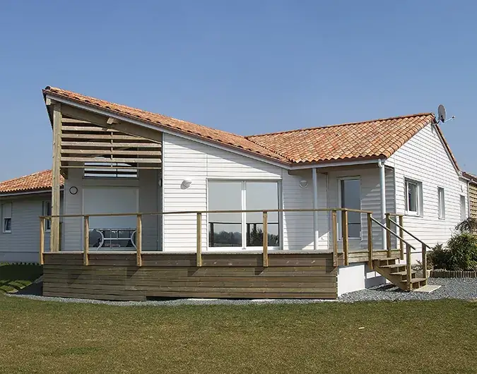 Maison traditionnelle en bois avec terrasse surélevée, toit en tuiles rouges et bardage blanc, sous un ciel dégagé.