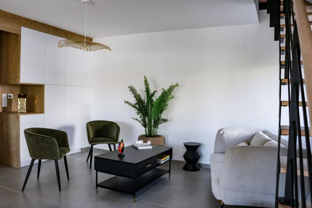 Salon moderne avec chaises vertes, table basse noire, canapé gris, plante décorative et escalier bois-métal, ambiance épurée.
