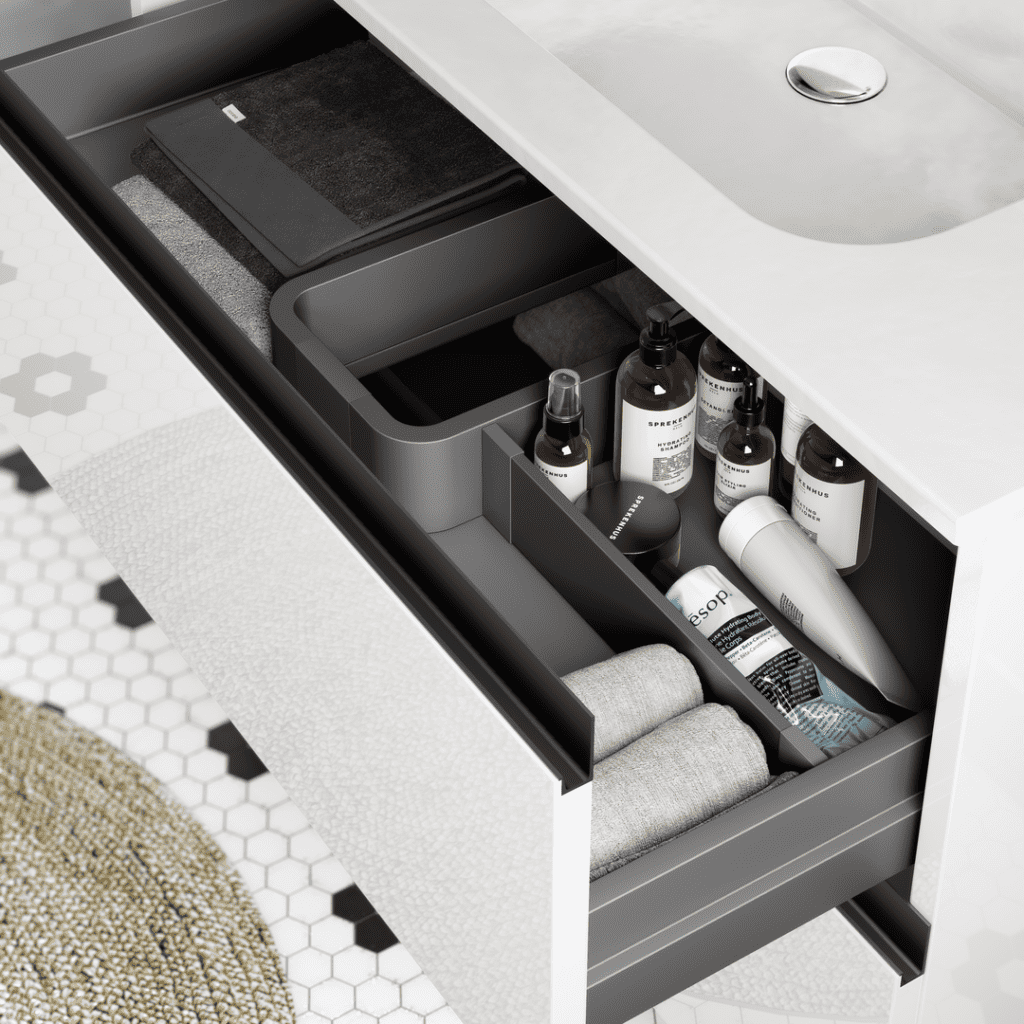 Un meuble de salle de bain moderne et fonctionnel, doté de tiroirs optimisés pour le rangement. Chaque espace est soigneusement organisé avec des produits de soin, des serviettes et accessoires bien disposés, offrant une vue claire sur l’intérieur. Le design épuré avec une finition blanc brillant s'harmonise avec le carrelage hexagonal et le tapis tissé, créant une ambiance élégante et pratique. Une solution idéale pour maximiser l'espace dans une salle de bain contemporaine.