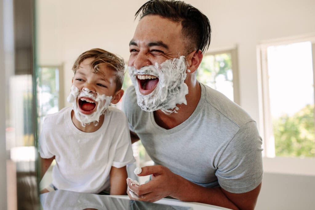 Une scène de complicité et de bonne humeur entre un père et son fils, illustrant un moment quotidien transformé en jeu. Tous deux partagent des éclats de rire en appliquant de la mousse à raser, symbolisant la joie des instants partagés en famille. Une lumière naturelle éclaire la pièce, renforçant l’ambiance chaleureuse et lumineuse.