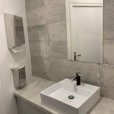 Salle de bain moderne avec carrelage texturé gris clair et un plan de travail en finition béton. La vasque rectangulaire blanche, associée à un robinet noir, apporte une touche élégante et contemporaine.