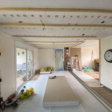 Rénovation d'une pièce en cours avec installation de rails de plafonds et préparation des murs. Les baies vitrées apportent une belle lumière naturelle, promettant un espace moderne et lumineux.