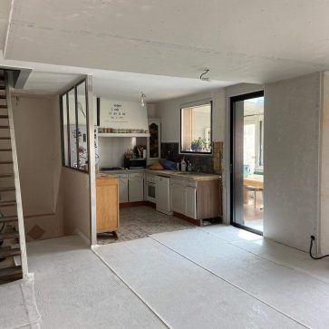 Intérieur d’une maison en pleine rénovation, intégrant une cuisine existante avec verrière et escalier en bois brut. L’espace en transformation promet une modernisation tout en conservant le charme des éléments existants.