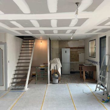 Travaux de rénovation en cours dans un espace ouvert avec cloisonnement et plafond en placo en phase de préparation. L'escalier en métal et bois ainsi que les grandes fenêtres promettent un intérieur lumineux et moderne.