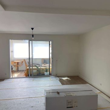 Pièce en cours d’aménagement avec un sol protégé, de grandes baies vitrées donnant sur une véranda lumineuse, et des murs prêts à accueillir la décoration. Un espace promettant un intérieur moderne et fonctionnel.