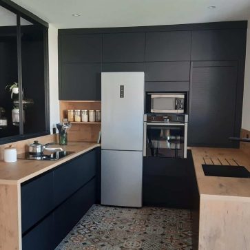 Cuisine moderne en U avec façades noires mates, plan de travail en bois clair, électroménager intégré et verrière industrielle. Le sol en carreaux de ciment ajoute une touche vintage à cet espace épuré et fonctionnel.