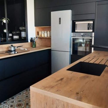 Cuisine moderne aux façades noires mates et plan de travail en bois clair, équipée d'un îlot central avec évier intégré. Le style épuré est rehaussé par une verrière industrielle et un sol en carreaux de ciment pour une ambiance chaleureuse et contemporaine.