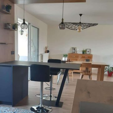 Vue d'une pièce à vivre moderne avec une cuisine ouverte équipée d'un îlot central noir et bois, des carreaux de ciment au sol, et une salle à manger lumineuse. Une ambiance conviviale et élégante alliant style contemporain et charme authentique.