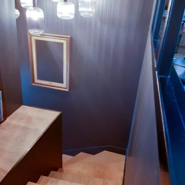 Escalier en bois clair éclairé par des suspensions en verre au design contemporain, avec des murs noirs et un miroir encadré doré pour une touche élégante et chaleureuse. Un espace moderne et raffiné.