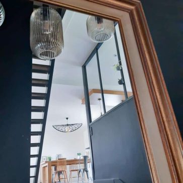 Reflet dans un miroir doré d’un intérieur moderne avec escalier en bois sombre, verrière industrielle et salle à manger lumineuse. Une combinaison élégante de matériaux et de styles pour un espace chaleureux et contemporain.