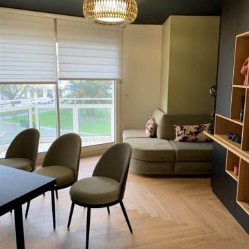 Salon moderne et chaleureux avec un parquet en chevrons, un canapé d’angle confortable et un coin repas élégant. La lumière naturelle met en valeur les teintes douces et les finitions soignées pour un espace convivial et raffiné.
