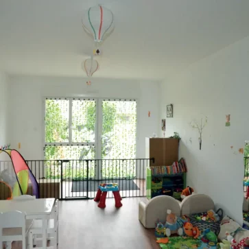 Cet espace a été transformé en une salle de jeux spacieuse et lumineuse dédiée aux enfants. Le parquet chaleureux et les murs blancs ornés de décorations enfantines créent une ambiance accueillante. L’agencement des zones de jeu, comprenant une tente colorée, des modules d’activités et des meubles adaptés, encourage la créativité et le développement. Les suspensions en forme de montgolfières apportent une touche poétique, tandis que les grandes baies vitrées laissent entrer une lumière naturelle généreuse, offrant une vue agréable sur l’extérieur.