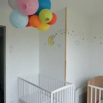 Cette pièce a été aménagée pour accueillir une chambre d'enfant lumineuse et chaleureuse. Les murs blancs, décorés de motifs colorés et de dessins enfantins, créent un univers apaisant et ludique. Les suspensions en papier multicolores ajoutent une touche joyeuse et festive, tandis que l'ouverture sur l'espace de vie apporte une belle continuité visuelle. Une réalisation qui combine fonctionnalité et esthétisme pour le bien-être des plus petits.