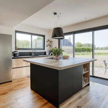 Cuisine moderne avec îlot central et grandes fenêtres offrant une vue sur la nature.