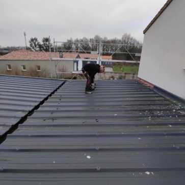 Travaux sur une toiture en bac acier avec un ouvrier en pleine intervention. L’image illustre le début d’un chantier d’extension ou de rénovation, mettant en avant la préparation et l’installation des éléments de couverture.