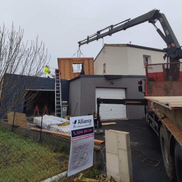 Chantier d'extension avec installation d'un mur en ossature bois à l'aide d'une grue. Un panneau est levé sur la plateforme préalablement posée, tandis que des ouvriers s'affairent à sécuriser la structure. Une signalisation de chantier et des matériaux sont visibles au premier plan, témoignant d'une organisation méthodique.