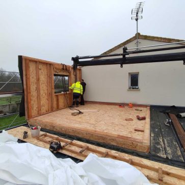 Chantier de surélévation en cours avec une ossature bois. Deux ouvriers positionnent un mur préfabriqué à l’aide d’une grue, mettant en avant la précision et la collaboration nécessaires pour l’installation. La plateforme en panneaux OSB est prête à accueillir les murs suivants.