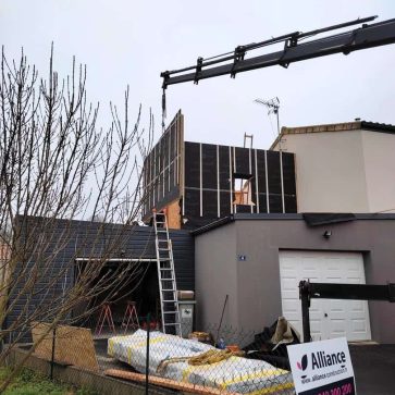 Chantier d’extension d’une maison avec une grue en action pour la mise en place des éléments de l'ossature bois. L’isolation extérieure est visible, ainsi que divers matériaux entreposés devant le garage. Une signalétique de chantier indique l'entreprise en charge des travaux.
