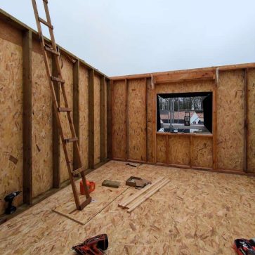 Intérieur d'une extension en construction, avec une ossature en bois et des panneaux OSB déjà installés. Une fenêtre en cours d’aménagement laisse apparaître l'extérieur, tandis que divers outils et matériaux de chantier sont disposés au sol, témoignant des étapes intermédiaires des travaux.