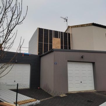 Extension en cours de réalisation sur une maison, avec une ossature bois recouverte d’un isolant noir et partiellement bardée en gris. L’élévation au-dessus du garage modernise la façade tout en optimisant les espaces. Les matériaux de construction sont visibles au premier plan.