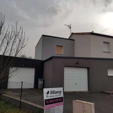 Maison en cours de rénovation avec une extension en bardage gris située au-dessus du garage. La façade moderne et l'ajout de l'étage témoignent d'une transformation contemporaine. Un panneau indique le chantier en cours