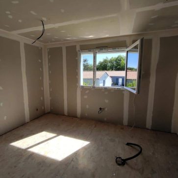 Chambre en cours de rénovation avec des murs en plaques de plâtre prêts à être peints et un sol en OSB. Une fenêtre ouverte laisse entrer la lumière naturelle, offrant une vue dégagée sur les maisons voisines. Les travaux électriques sont en préparation pour un espace moderne et fonctionnel.
