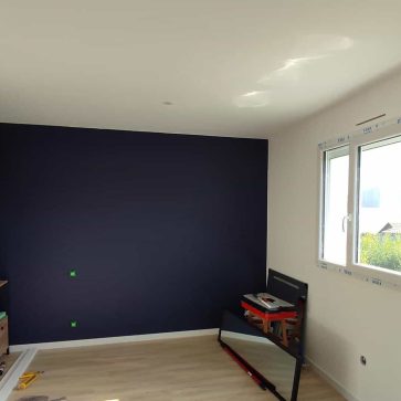Chambre rénovée avec un mur peint en bleu profond contrastant avec des murs blancs et un sol en parquet clair. Les travaux de finition sont en cours, avec des outils et des meubles partiellement installés, annonçant une ambiance moderne et chaleureuse.