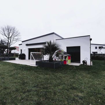 Extérieur d'une maison rénovée dans un style contemporain, avec une terrasse spacieuse, des espaces verts bien entretenus, et des équipements pour enfants, tels qu'une balançoire et un toboggan, soulignant un aménagement familial et moderne.