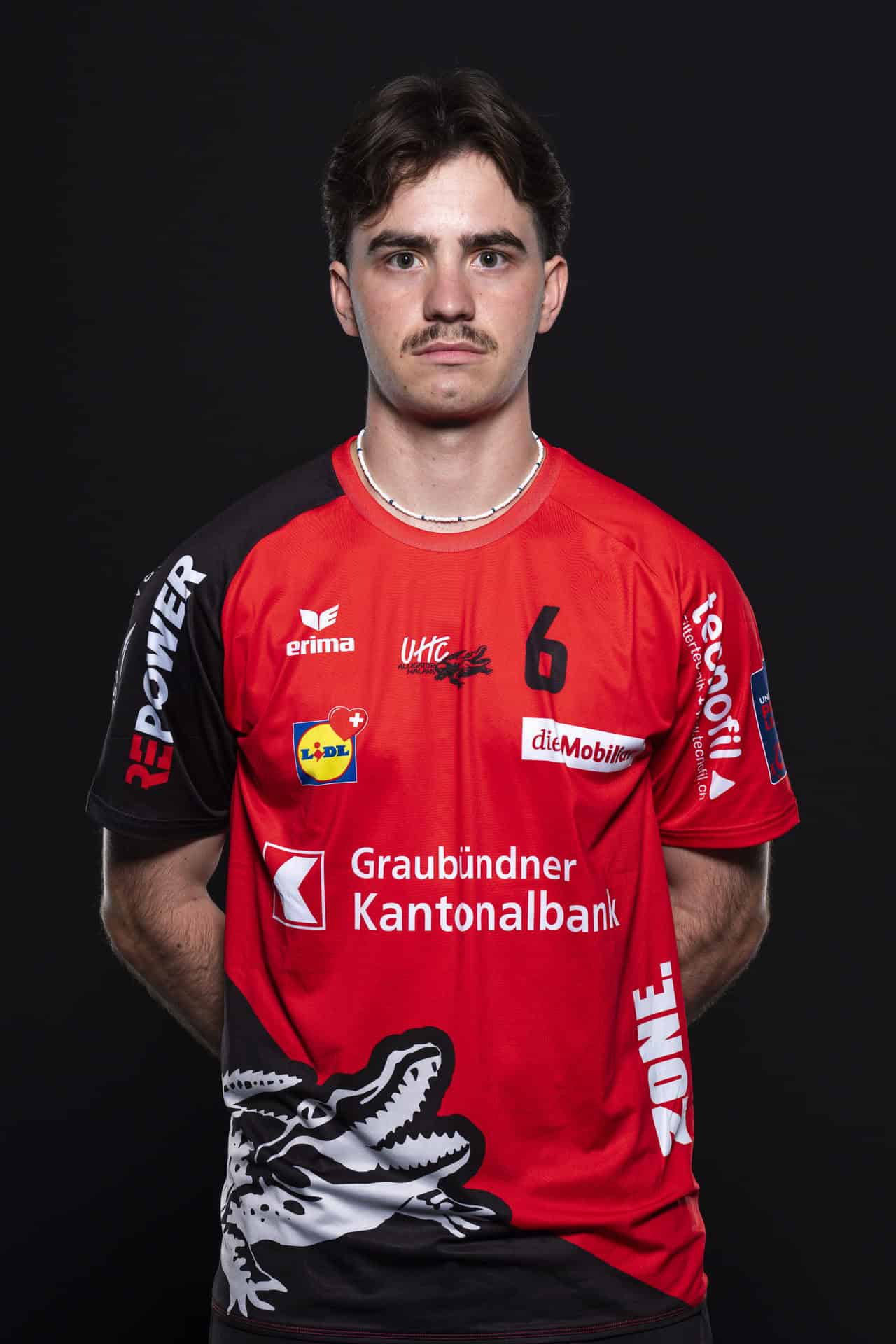 UHC Alligator Malans Volleyballspieler im Trikot, professionelle Sportfotografie, Blick in die Kamera, Teamfarben Rot und Schwarz, Sponsorenlogos sichtbar, regionaler Sportverein, sportliche Atmosphäre.