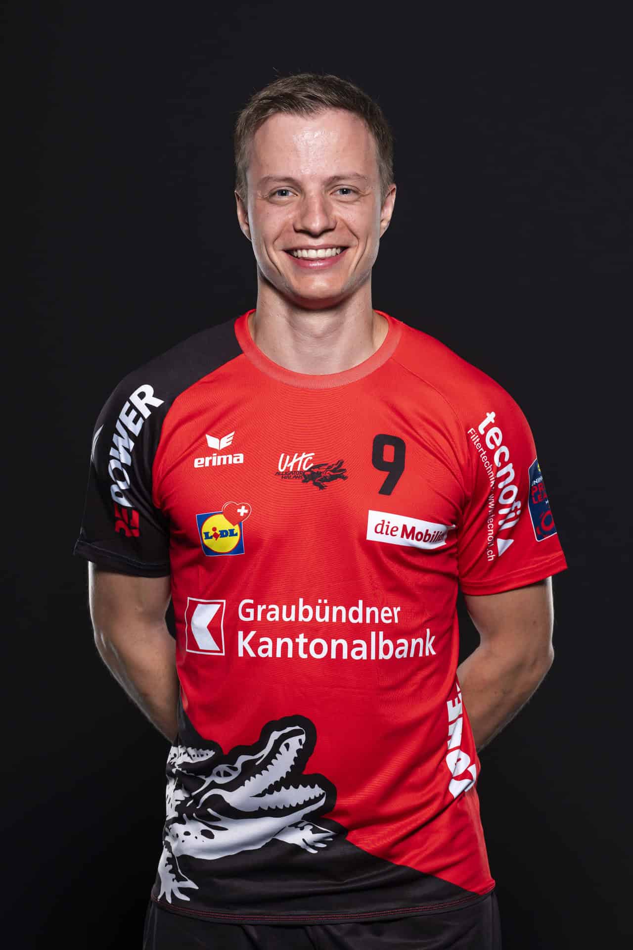 Hochwertiger Sportler in rotem Volleyball-Trikot mit Nummer 9, lächelnd vor schwarzem Hintergrund, präsentiert Sponsorenlogos und den Markenaufdruck von UHC Alligator Malans.
