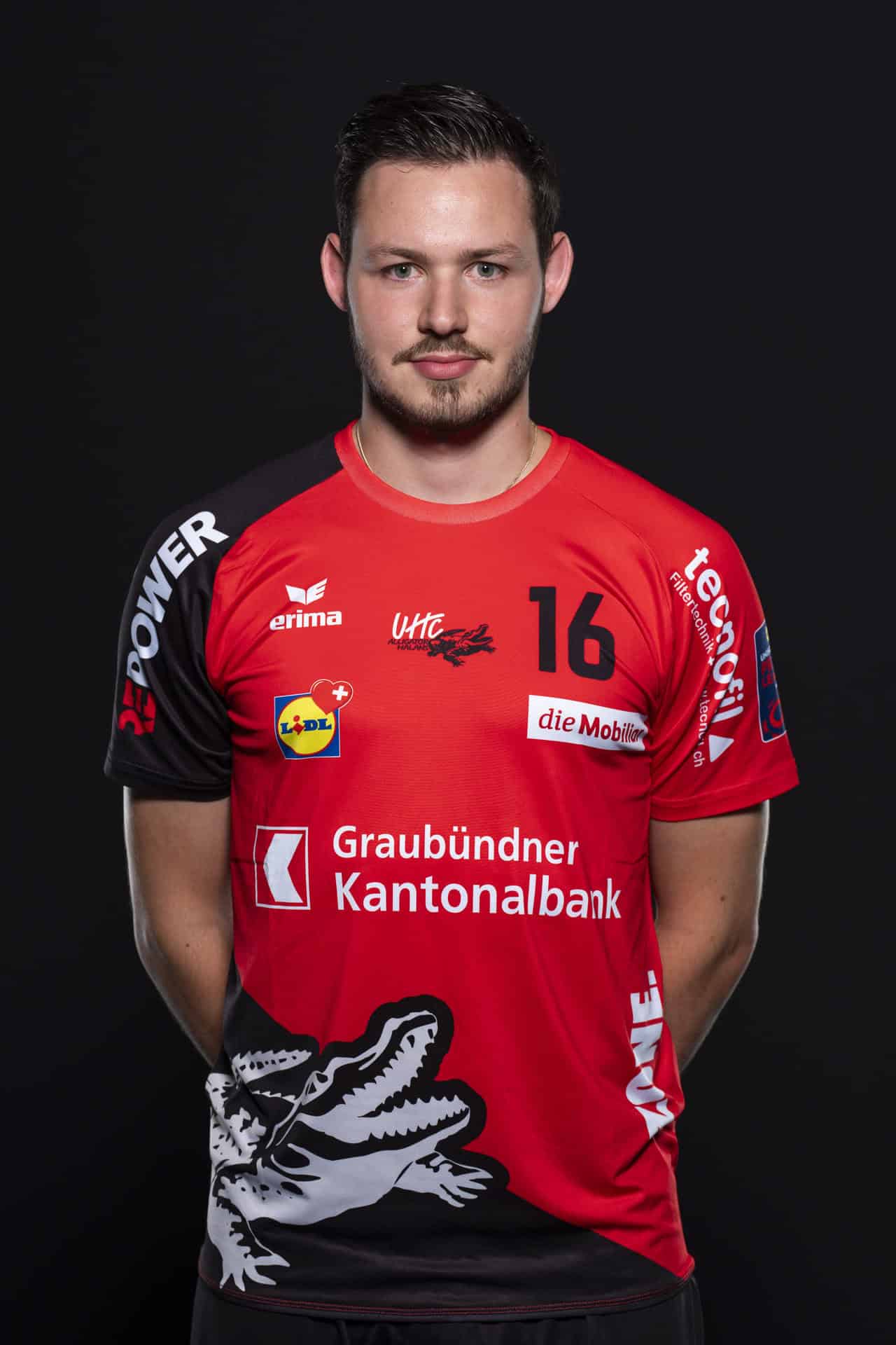 Hochwertiges Teamfoto eines Handballspielers in Rot vor schwarzem Hintergrund, trägt UHC Alligator Malans Trikot mit Sponsoren und Nummer 16, symbolisiert Engagement im Schweizer Handball.