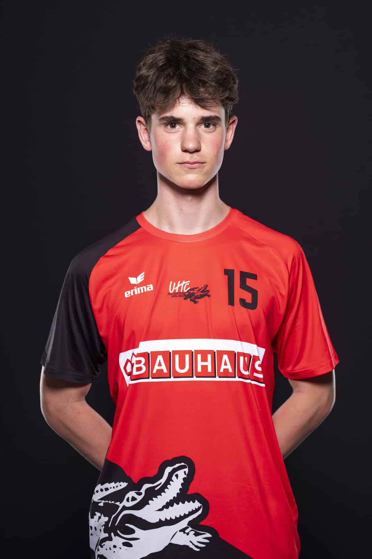 Jugendlicher Handballspieler in Rot- und Schwarztrikot von UHC Alligator Malans, Frontansicht gegen dunklen Hintergrund, zeigt Stolz und Teamgeist, symbolisiert Engagement im Handballsport.