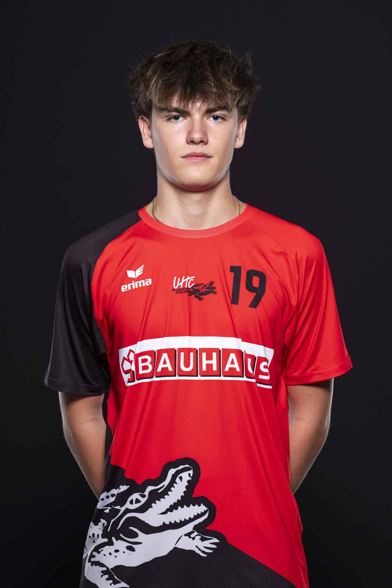 UHC Alligator Malans Handballspieler in rotem Trikot mit schwarzem Ärmel, lässig und professionell gestylt, steht vor schwarzem Hintergrund.