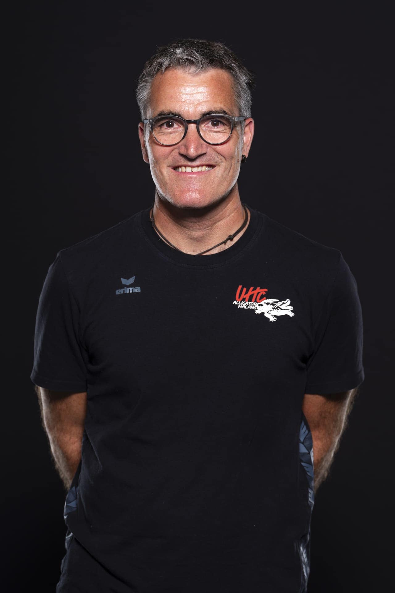 Sportlerportrait des Trainers des UHC Alligator Malans, lächelnd, in Vereinsmannschafts-Shirt, Schwarz Hintergrund, Fokus auf Gesicht und Team-Logo, Ausdruck von Freundlichkeit und Teamgeist.