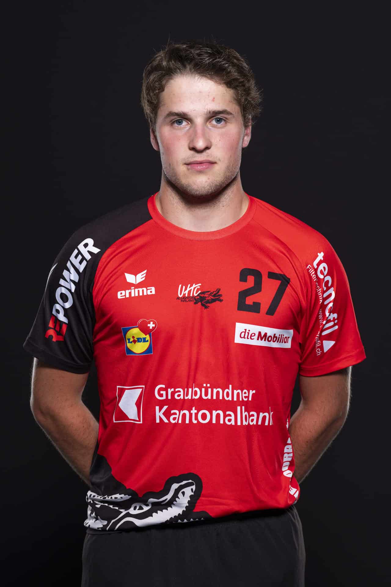 Sportlerporträt des UHC Alligator Malans in rotem Trikot mit Sponsoren, Nummer 27, auf dunklem Hintergrund.