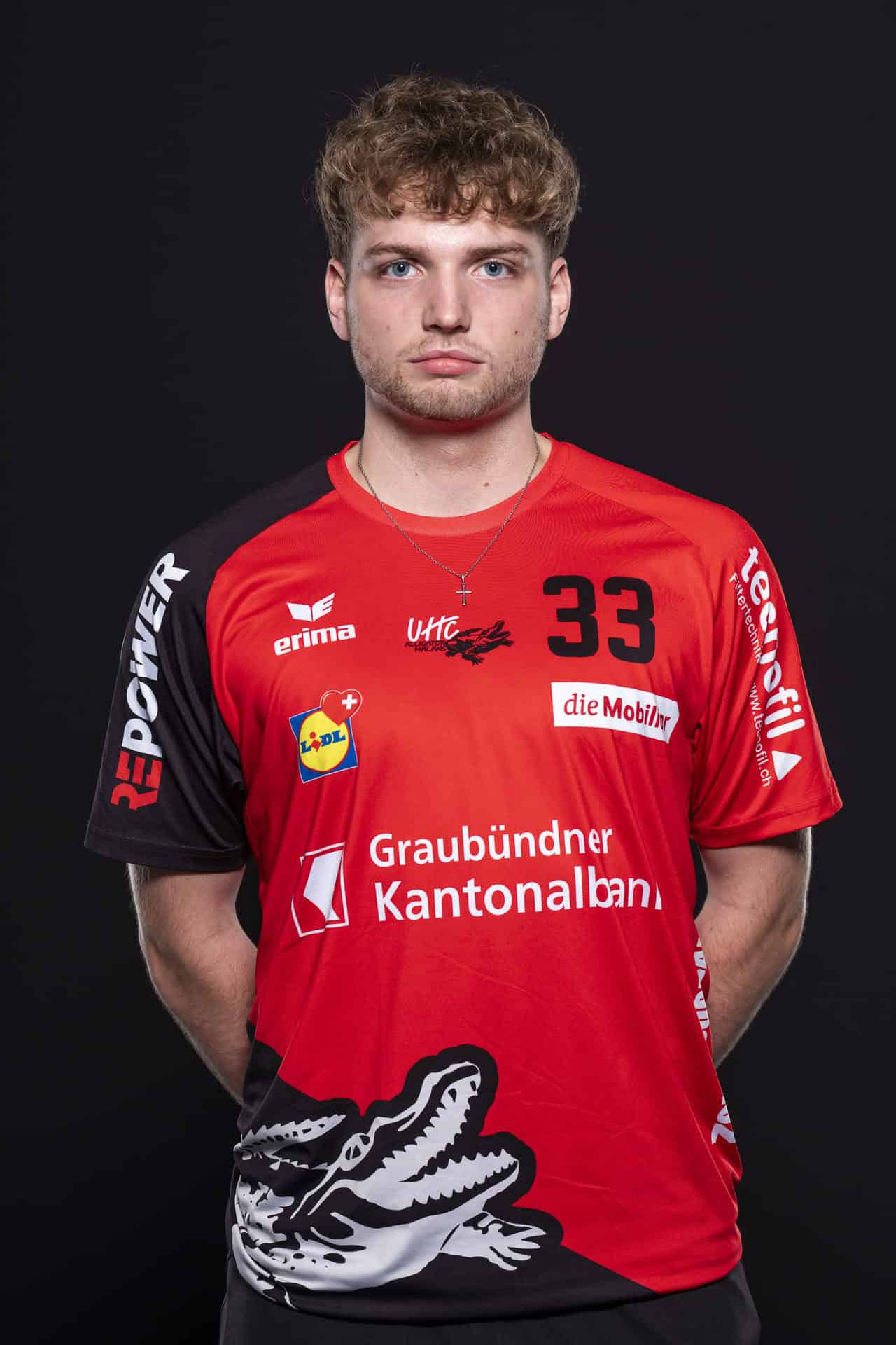 Hochwertiges Mannschaftsporträt eines jungen Handballspielers vom UHC Alligator Malans, in rotem Trikot mit Sponsorenlogos, gegen schwarzen Hintergrund.