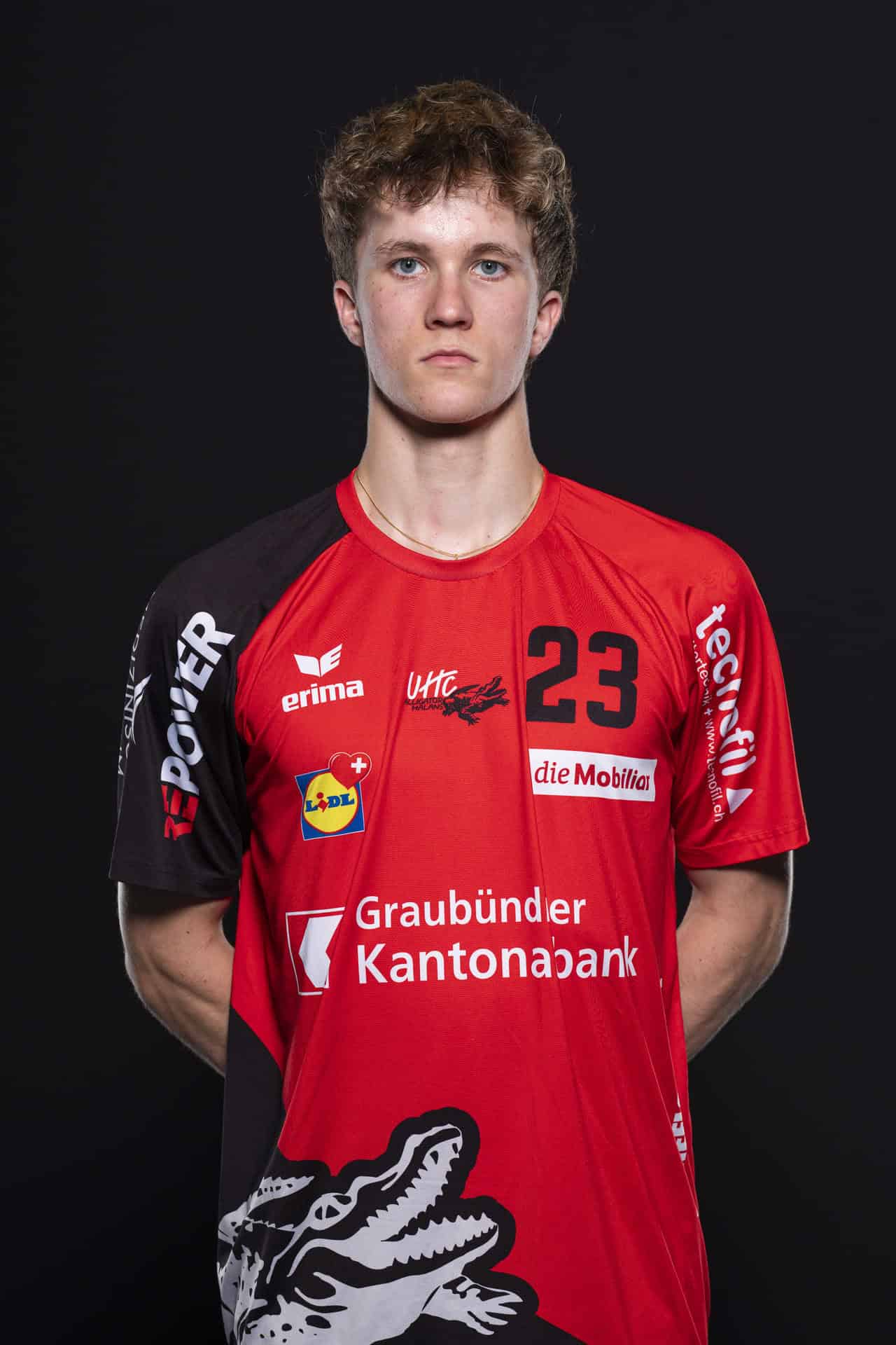 UHC Alligator Malans Handballspieler in rotem Trikot, stolz und fokussiert, mit Nummer 23, im professionellen Teamportrait vor schwarzem Hintergrund.
