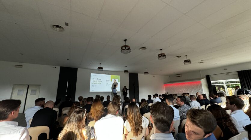 Présentation devant public, salle tamisée, présentateur debout, écran projetant du texte.