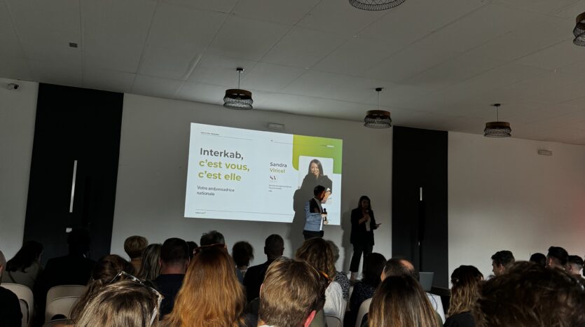Personnes assises regardant une présentation sur scène dans une salle de conférence bien éclairée.