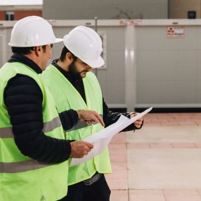 Dos ingenieros con cascos y chalecos reflectantes revisando planos en obra de infraestructura eléctr.